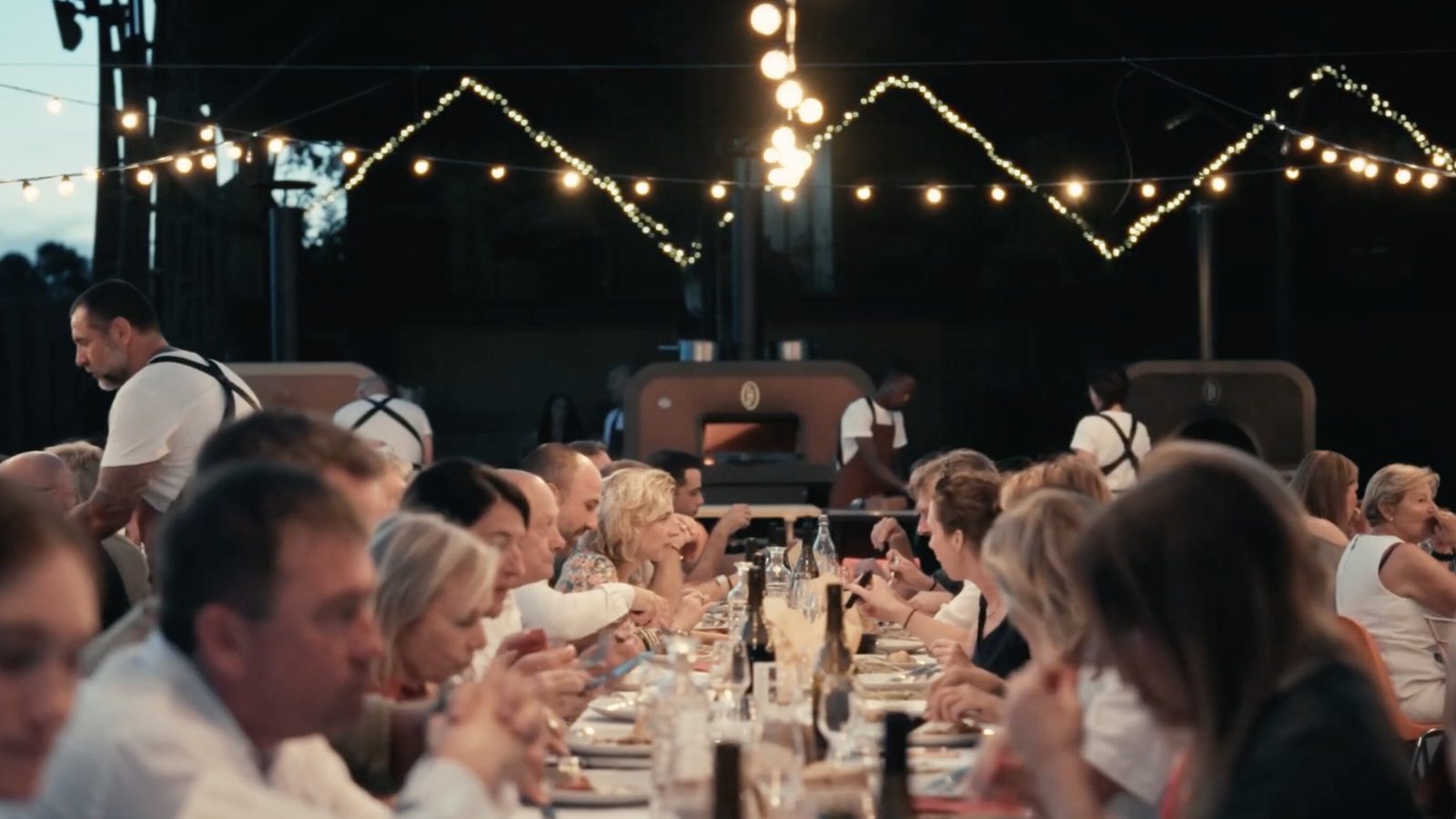 A long table at dusk under festoon lights, Le Flambeur ovens cooking behind